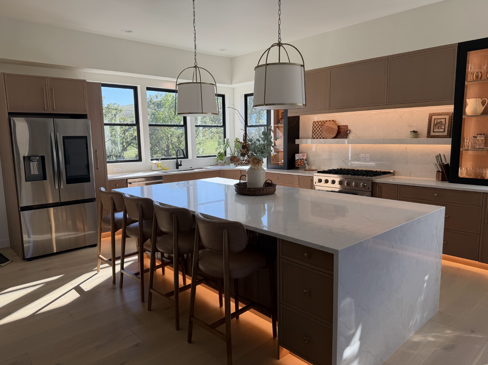 Kitchen with light oak cabinetry, marble waterfall island, brass lantern pendants, floating marble shelves and a glowing under cabinet light wash