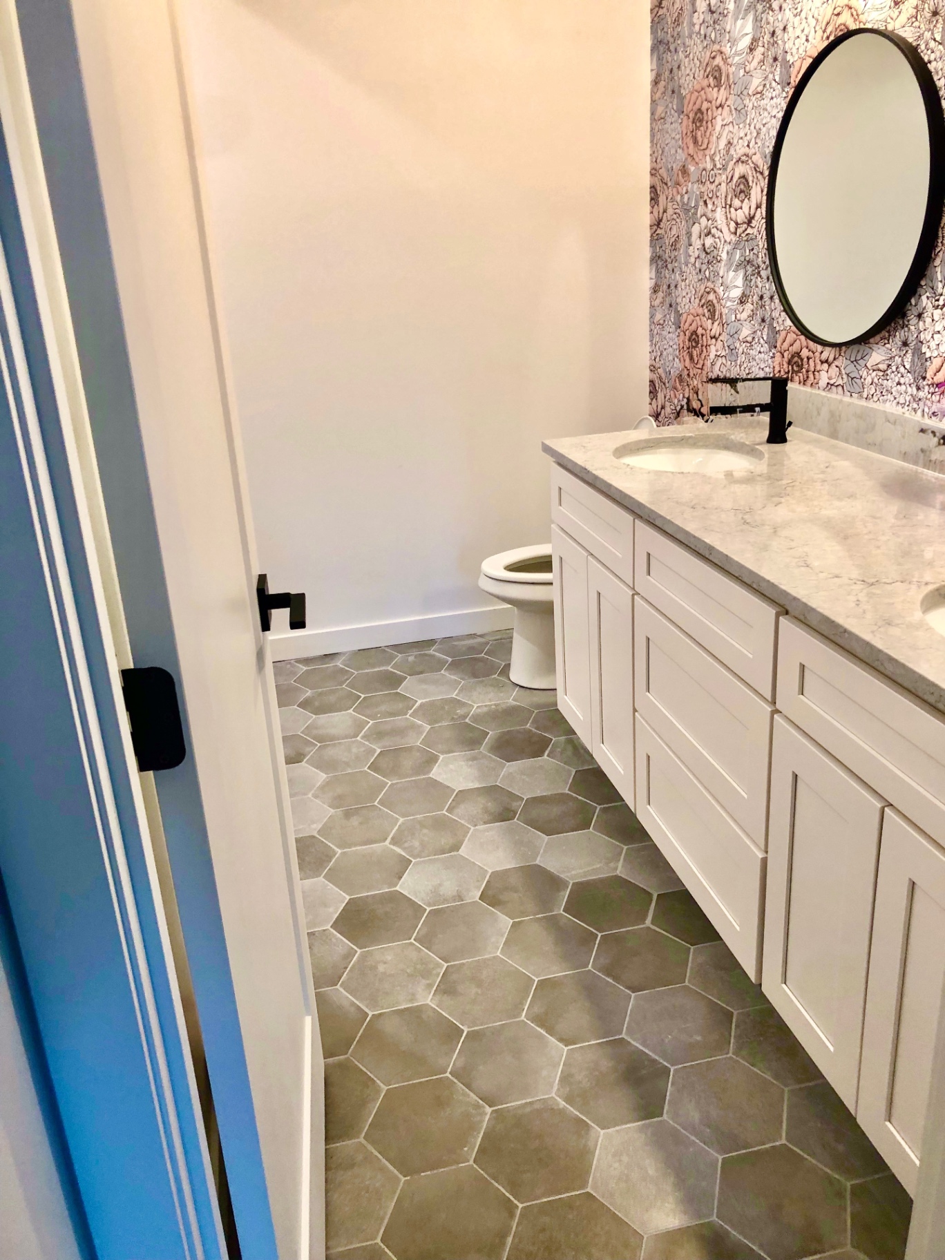 Powder bath with floral wallpaper, white shaker double vanity, quartz top and round black mirror