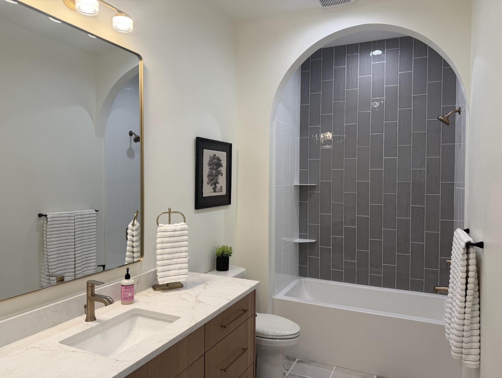 Bathroom with white oak double vanity, brass faucet, marble counter and a tub and shower with linear wood look tile in an arched opening